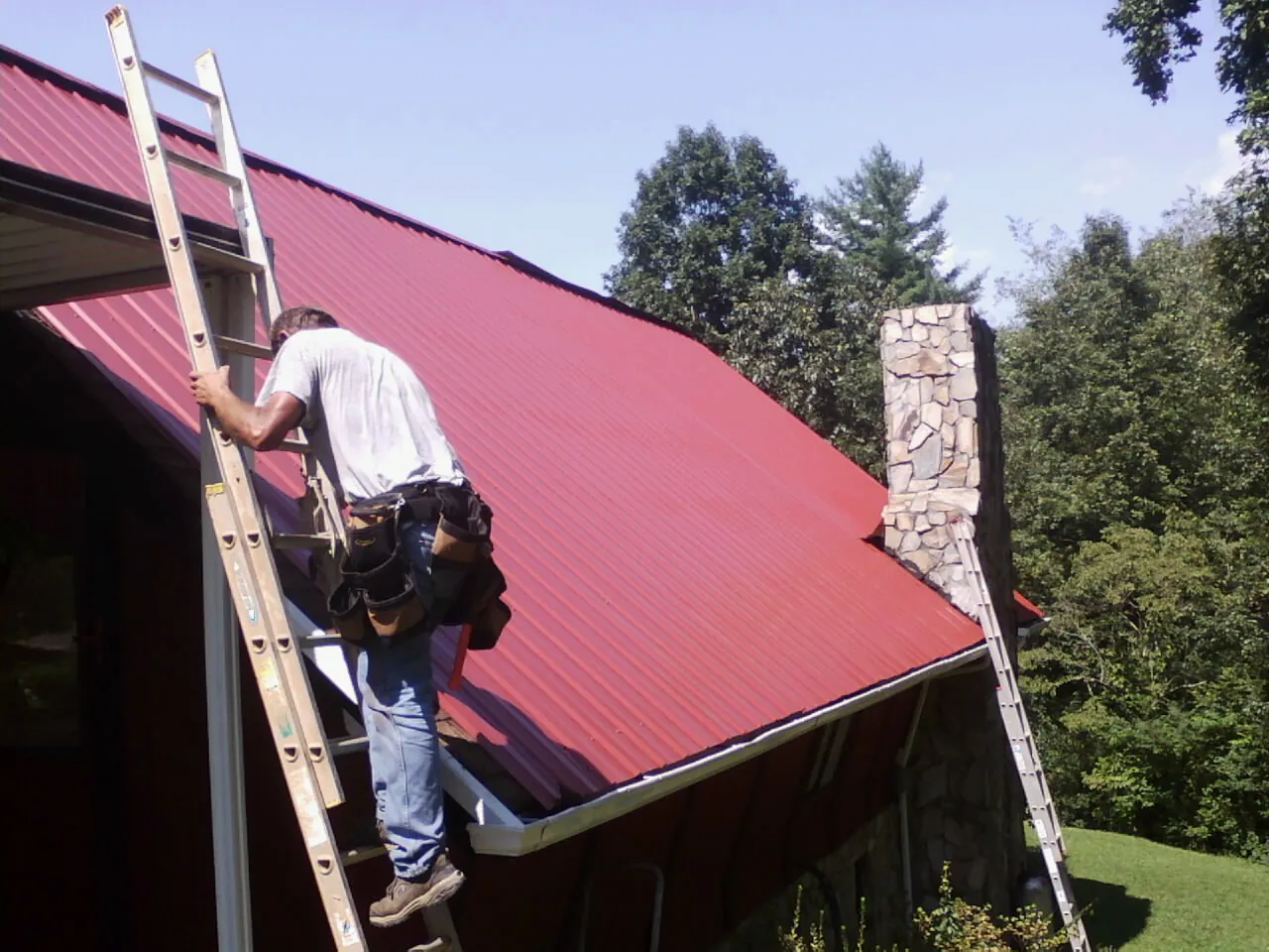 Roofer on a ladder beside a red metal roof and stone chimney — Soffit & Fascia Repair by Roofing Brothers in Birmingham